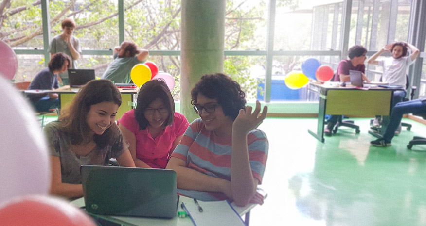 Estudantes lendo em um notebook em um sala bem iluminada, com árvores no fundo