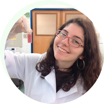 Aluna vestida com um jaleco no laboratório de ciências, segurando um pequeno vidro preenchido com um líquido avermelhado em sua mão esquerda.