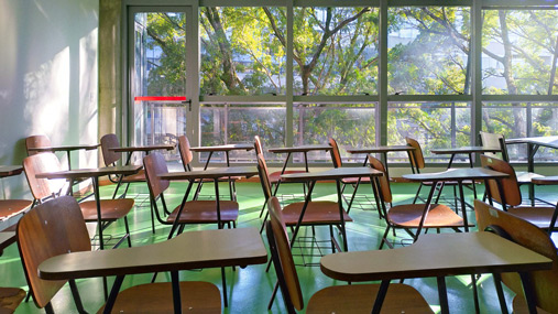 Sala de aula bem iluminada com amplas janelas de vidro, onde é vista uma paisagem arborizada.